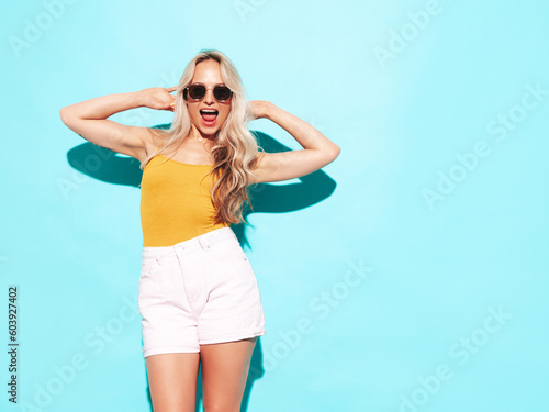 Young beautiful smiling blond female in trendy summer shorts clothes. Sexy carefree woman posing near blue wall in studio. Positive model having fun. Cheerful and happy. Going crazy