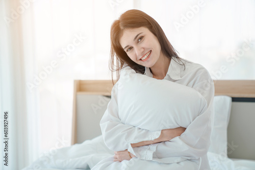 Photo of young happy woman in pajama stretching her arms and smiling while sitting on bed after sleep