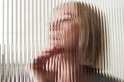 Horizontal through textured glass wall portrait of pensive senior Caucasian woman with blond bob cut hair, striped distortion effect photo