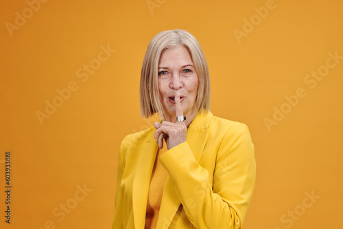 Horizontal medium portrait of attractive senior woman wearing yellow outfit saying hush or be quiet, bright yellow background photo