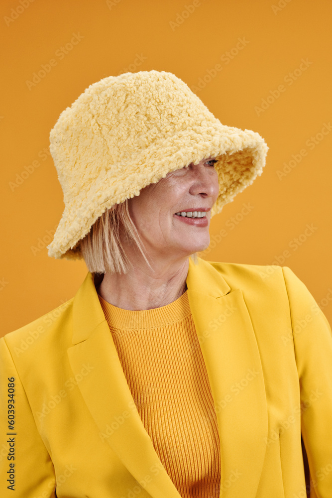 © Clique Images - Vertical medium close-up portrait of modern mature Caucasian woman wearing stylish yellow outfit with hat looking away, bright yellow background