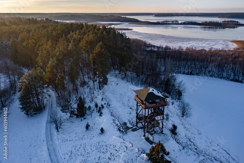 Winter drone landscape - observation tower, wooden house near forest, frozen Wigry lake in the background, Poland, Suwalszczyzna