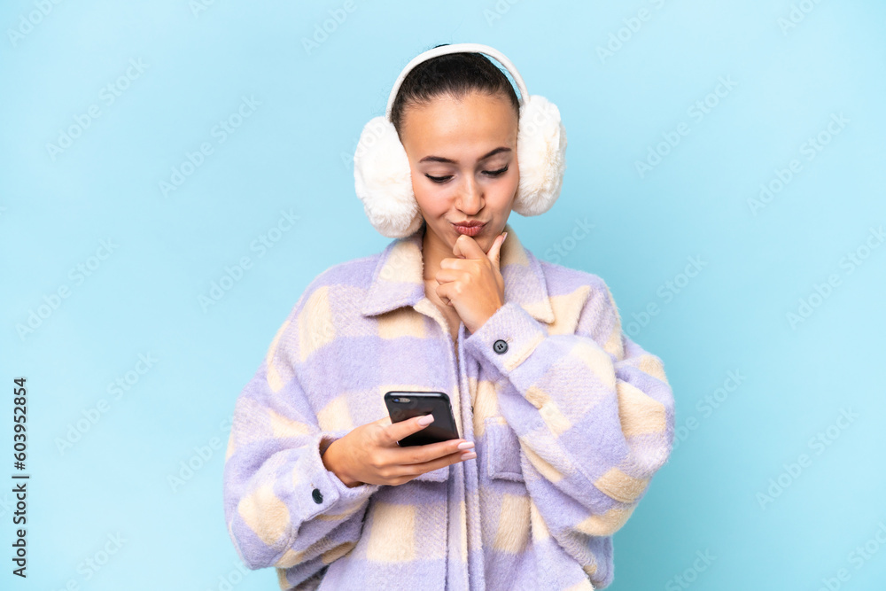 Young Arab woman wearing winter muffs isolated on blue background thinking and sending a message Young Arab woman wearing winter muffs isolated on blue background thinking and sending a message