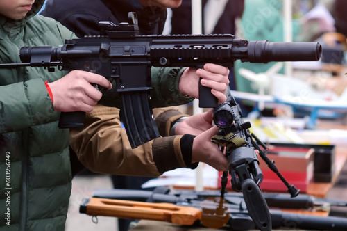 Assault rifle in the hands of a teenager at an arms exhibition. Inspection of automatic small arms. Recruitment of new recruits for military service. Army, ammunition, war, danger concept.