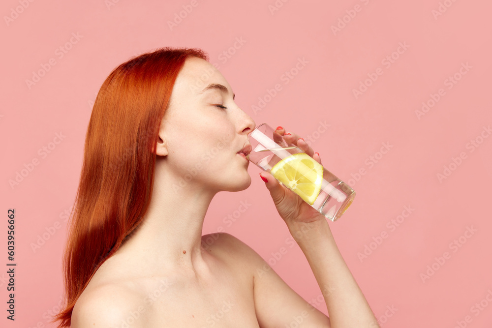 Beautiful girl with perfect skin and red hair holding glass of water and drinking with closed eyes over pink background. Side view. Water balance Beautiful girl with perfect skin and red hair holding glass of water and drinking with closed eyes over pink background. Side view. Water balance
