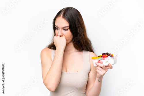 Young caucasian woman holding a bowl of fruit isolated on white background having doubts