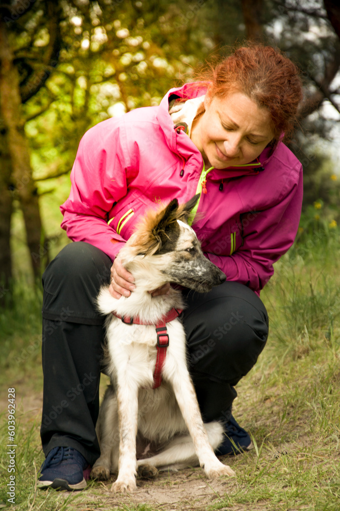 Woman hugging dog in forest. Cheerful lady in pink jacket hugs puppy sitting on a path of the forest on nice day. Woman hugging dog in forest. Cheerful lady in pink jacket hugs puppy sitting on a path of the forest on nice day.
