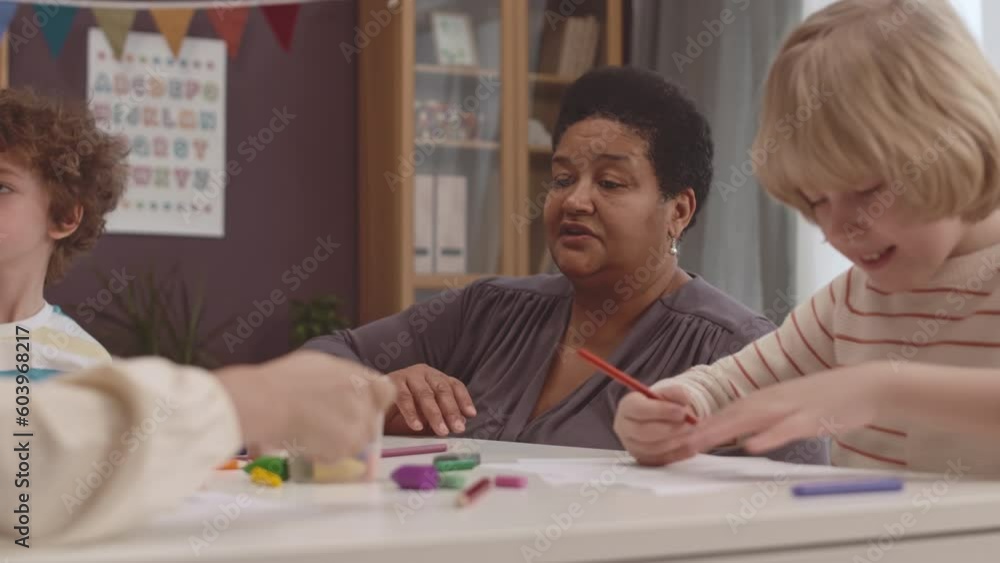 Chest up of mature Black female teacher working with group of elementary age kids drawing pictures in preschool Chest up of mature Black female teacher working with group of elementary age kids drawing pictures in preschool