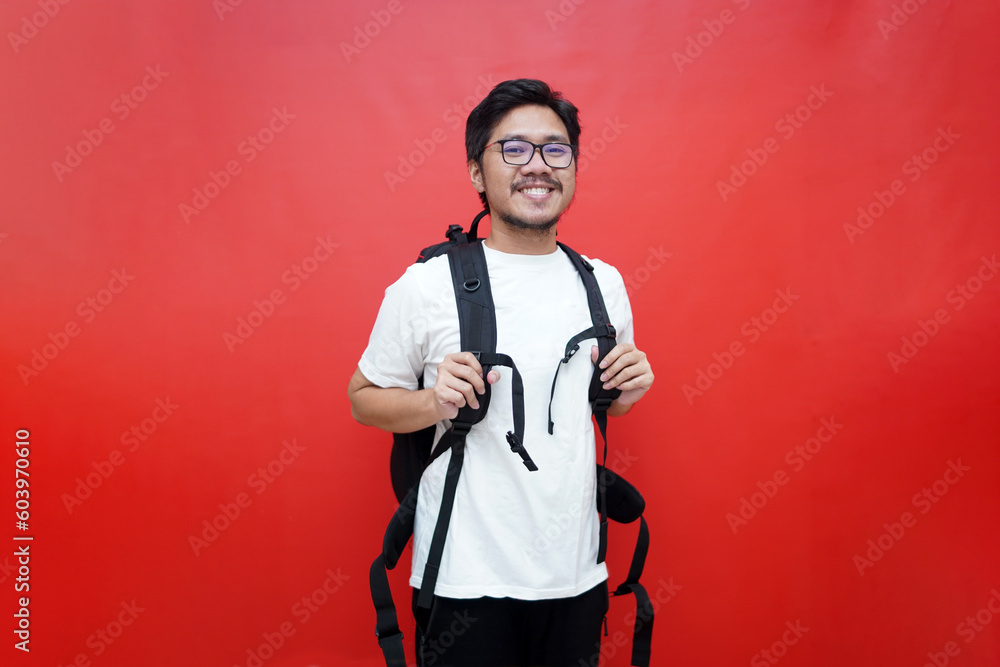 Portrait of smiling young Asian tourist male carrying backpack ready to travel in studio isolated red background Portrait of smiling young Asian tourist male carrying backpack ready to travel in studio isolated red background