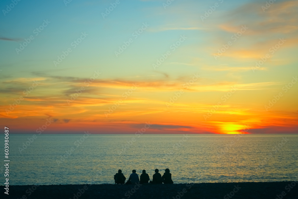 Young teenage people - guys and girls - sit on the beach and watch the sunset. Group of five happy people sits on background of empty sunset beach. Travel or sea vacations concept. back, rear view.