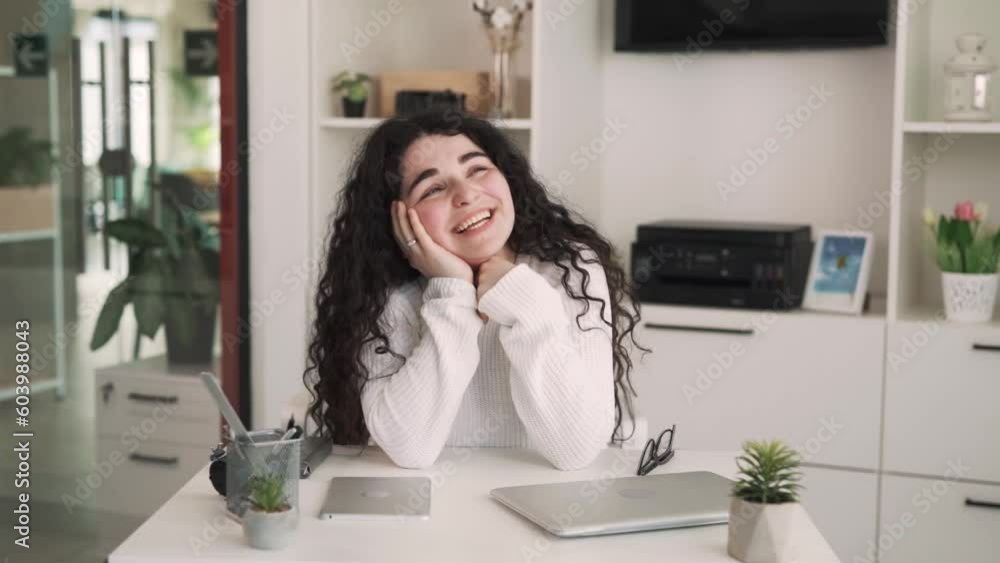 The young brunette woman with long curly hair is sitting at the table, resting her chin on her hand and daydreaming. She smiles, imagining something pleasant. The young brunette woman with long curly hair is sitting at the table, resting her chin on her hand and daydreaming. She smiles, imagining something pleasant.