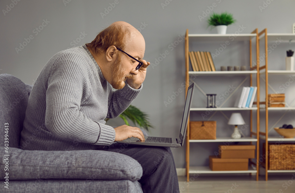 Elderly man touching his glasses rim looking attentively at laptop computer. Side view portrait of bearded elderly man learning to use modern device, mastering internet or reading news