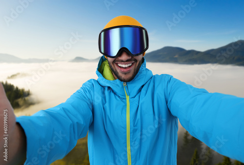 Smiling young man in ski goggles taking selfie in mountains