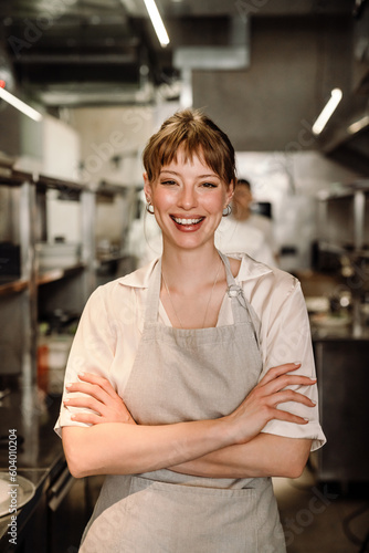 Cheerful young blonde woman business owner in apron looking at camera in cafe kitchen