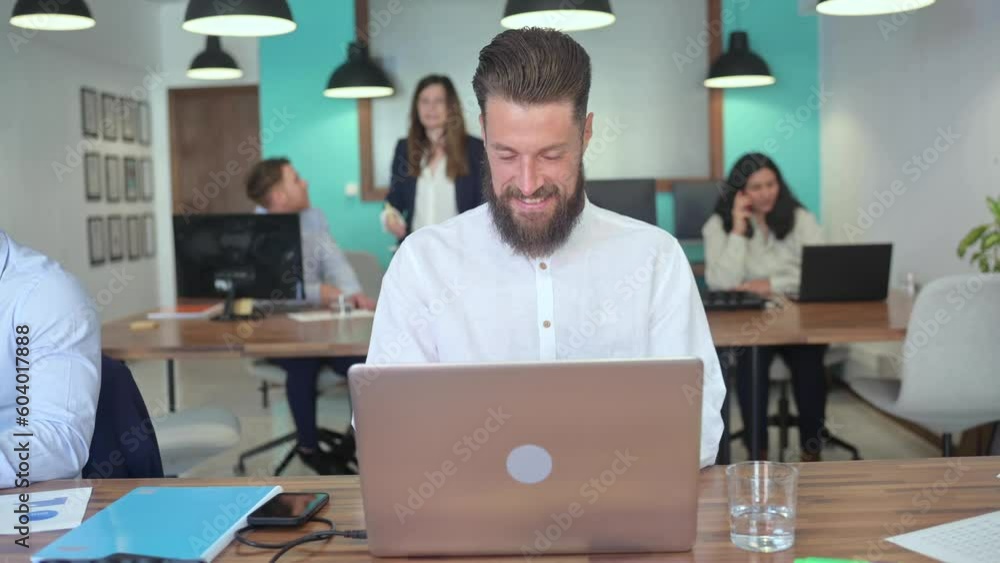 Real time of young positive bearded male office worker sitting at table against colleagues and working on netbook in modern workspace during workday Real time of young positive bearded male office worker sitting at table against colleagues and working on netbook in modern workspace during workday