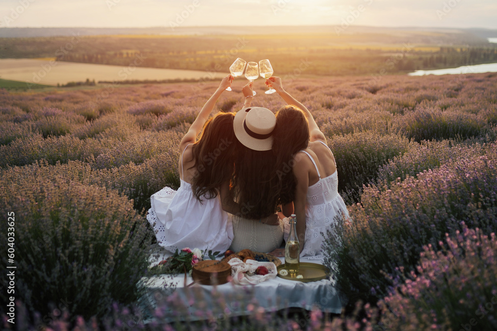 Young women enjoying view of sunset in the lavender field. They clinking glasses of wine to celebrate their vacation.