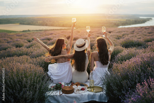 Young female friends having fun, raising glasses with wine and enjoy beautiful sunset at summer picnic in lavender field Fototapet
