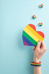 Overhead first person view vertical shot of a young woman's hand wearing a symbolic wristlet, holding a pride flag card over badges on a pastel blue background with empty space for a message or ad