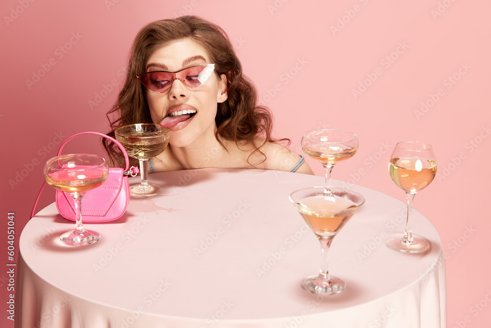 Portrait of young beautiful woman peeking out table served with champagne against pink studio background. Funny crazy mood. Concept of emotions, celebration, party, fashion, beauty Portrait of young beautiful woman peeking out table served with champagne against pink studio background. Funny crazy mood. Concept of emotions, celebration, party, fashion, beauty
