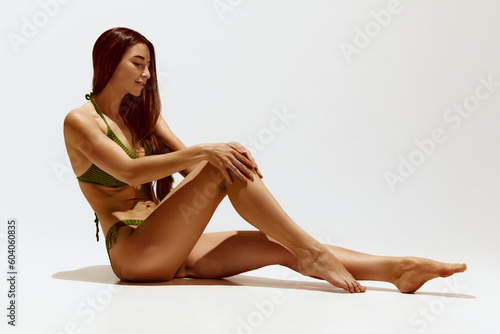 Portrait of tender, beautiful young woman in green swimsuit with tanned, smooth, slim body sitting, posing against grey studio background. Concept of body, skin care, spf protection, cosmetics, beauty photo