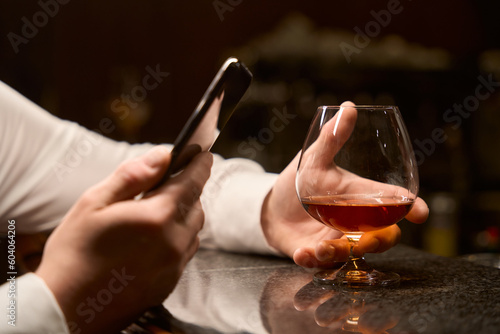 Male hands holding smartphone and glass of whiskey at bar counter