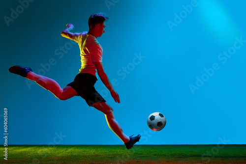 Professional man, football player in sports team uniform running over soccer field background in neon light. Strong kick