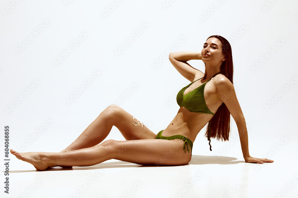Portrait of beautiful young woman with tanned body in swimsuit, applying spf cream against white studio background. Wellness. Concept of body, skin care, spf protection, cosmetics, beauty Portrait of beautiful young woman with tanned body in swimsuit, applying spf cream against white studio background. Wellness. Concept of body, skin care, spf protection, cosmetics, beauty