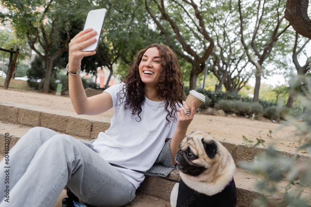 Cheerful young and curly woman in casual clothes having video call on smartphone and holding takeaway coffee near pug dog sitting on stairs in blurred park at daytime in Barcelona, Spain