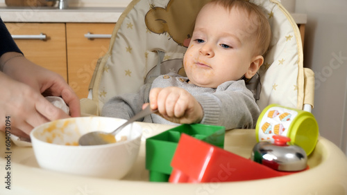 Portrait of mother wiping with paper towel her messy baby boy eating porridge in highchair. Concept of parenting, healthy nutrition and baby feeding.