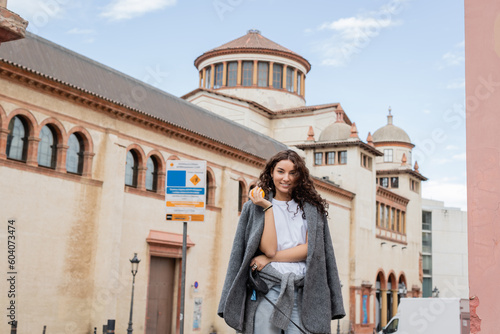 Smiling and brunette woman in casual jacket holding fresh orange and leash while standing near blurred historic landmark on urban street in Barcelona, Spain, ancient building