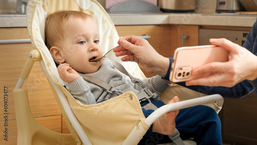 Mother showing video on smartphone while feeding her baby in highchair. Concept of parenting, healthy nutrition and baby feeding.