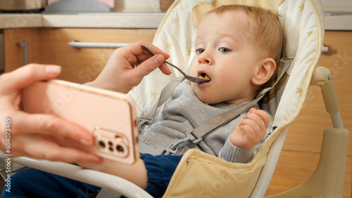 Portrait of cute baby boy eating porridge in highchair and watching cartoons on smartphone