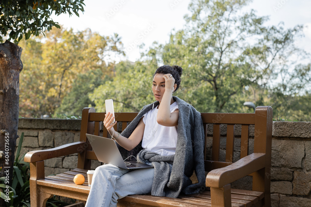 Fototapeta premium Curly and pretty freelancer in warm jacket having video call on smartphone near laptop, coffee to go and orange on wooden bench in park at daytime in Barcelona, Spain