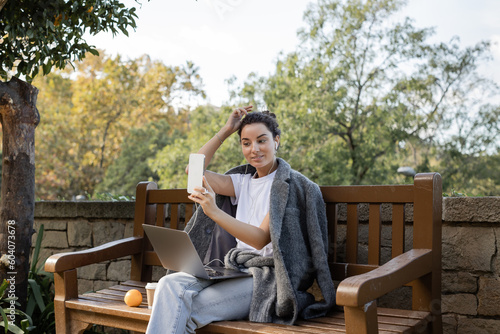 Smiling and young freelancer in warm jacket using earphones and smartphone while sitting near laptop, takeaway drink and fresh orange on wooden bench in park in Barcelona, Spain