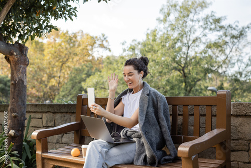 Foto Cheerful young freelancer in warm jacket and earphones waving hand at smartphone