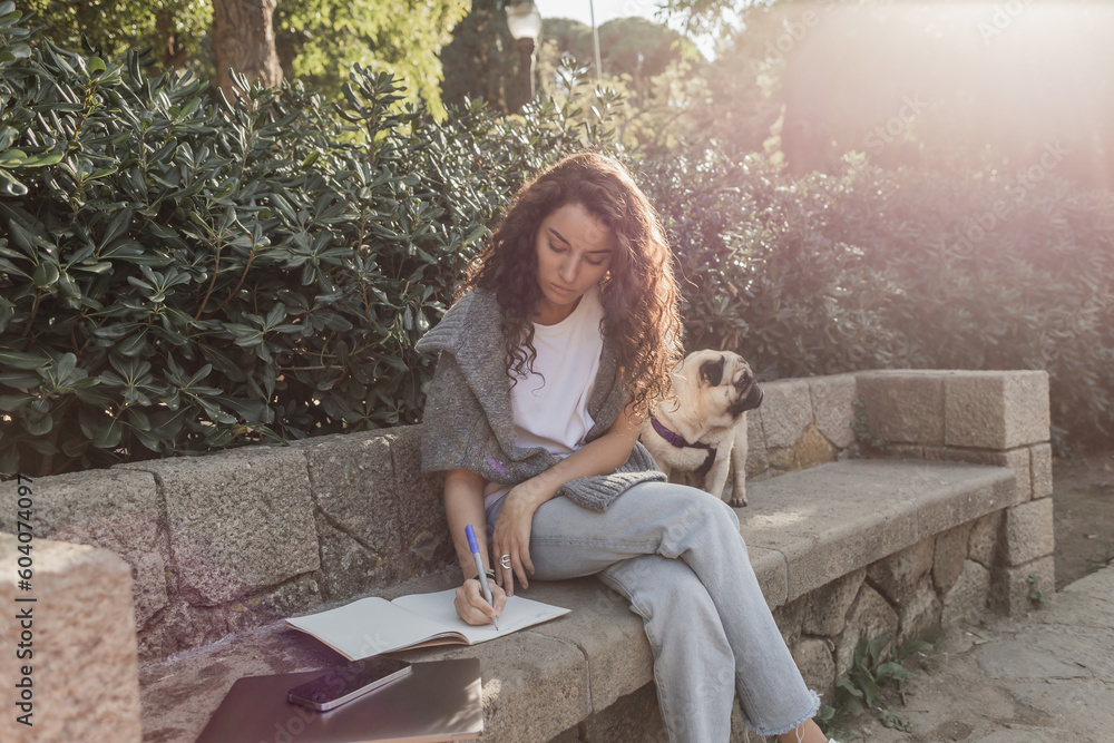 Curly and young woman in casual clothes writing on notebook while sitting near gadgets and pug dog on stone bench and green bushes in park in Barcelona, Spain, freelance Curly and young woman in casual clothes writing on notebook while sitting near gadgets and pug dog on stone bench and green bushes in park in Barcelona, Spain, freelance