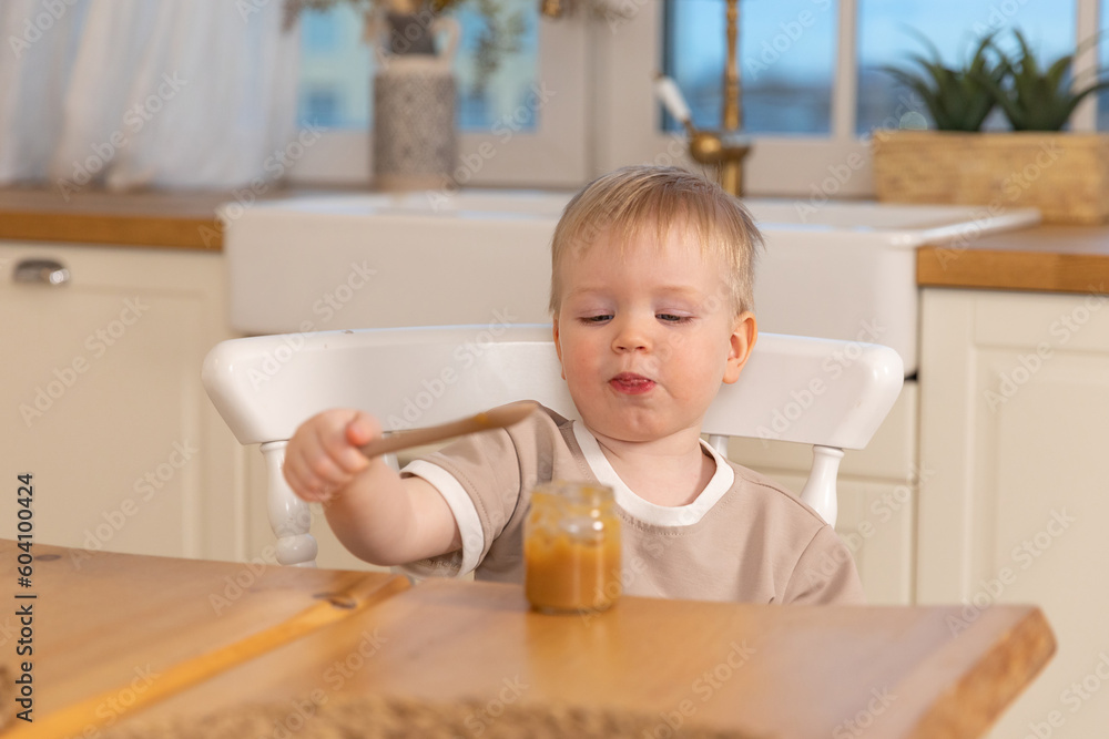 Happy family at home. Baby boy feeding himself in kitchen. Little boy with messy funny face eats healthy food. Child learns eat by himself holding spoon. Self feeding Happy family at home. Baby boy feeding himself in kitchen. Little boy with messy funny face eats healthy food. Child learns eat by himself holding spoon. Self feeding