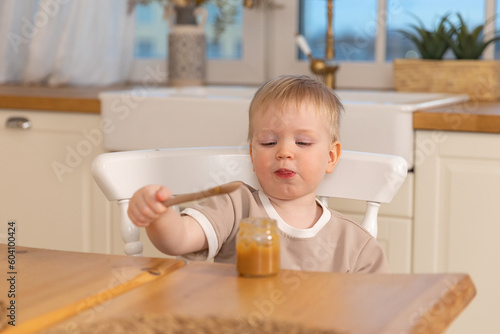 Happy family at home. Baby boy feeding himself in kitchen. Little boy with messy funny face eats healthy food. Child learns eat by himself holding spoon. Self feeding