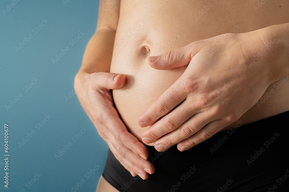 Very close up view of pregnant woman holding her belly gently during first months of pregnancy. Pregnancy first trimester - week 18. Side angle view. Blue background. Bright shot. Very close up view of pregnant woman holding her belly gently during first months of pregnancy. Pregnancy first trimester - week 18. Side angle view. Blue background. Bright shot.