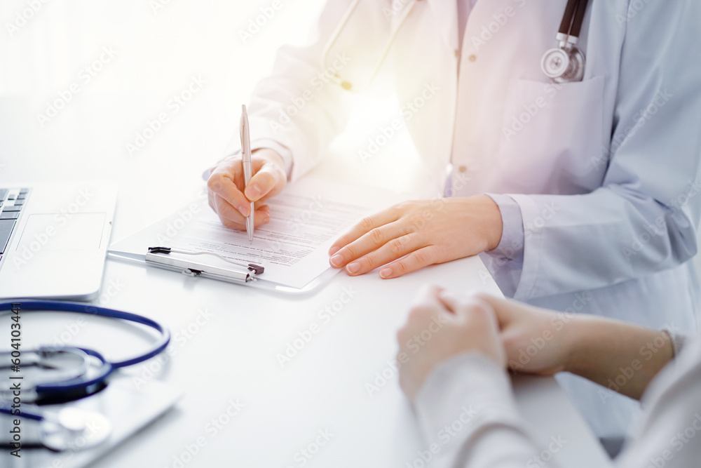 Doctor and patient sitting at the table in clinic. The focus is on female physician's hands filling up the medication history record form or checklist, close up. Medicine concept. Doctor and patient sitting at the table in clinic. The focus is on female physician's hands filling up the medication history record form or checklist, close up. Medicine concept.