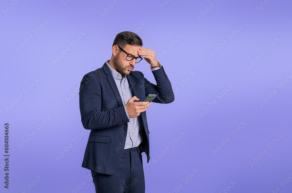 Upset and worried young businessman touching head while reading bad news over smart phone. Stressed male manager checking his messages on cellphone while standing against blue background Upset and worried young businessman touching head while reading bad news over smart phone. Stressed male manager checking his messages on cellphone while standing against blue background