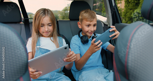 Beautiful children boy and girl having fun playing games on phone and tablet while sitting in a modern car. Little brother and sister are laughing and happily celebrating vin in online game by giving © serg