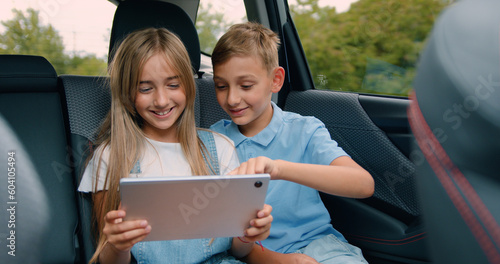 Handsome little boy in blue shirt is taking selfie with smartphone while sitting on the back seat of car. Fun family trip.