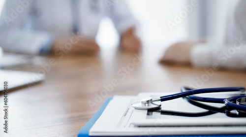 Stethoscope and tablet computer are lying on the wooden table while doctor and patient discussing something at the background. Medicine concept.