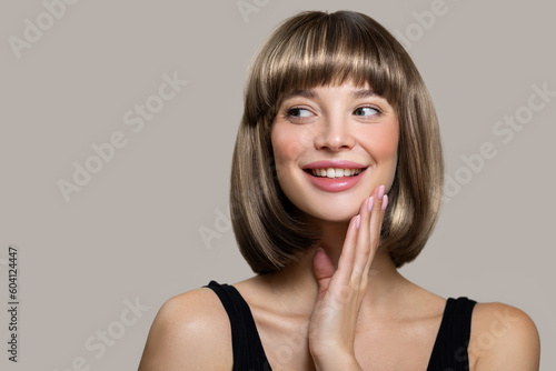 Happy young woman with bob hairstyle smiling while touching her face with her hand. Gray background photo