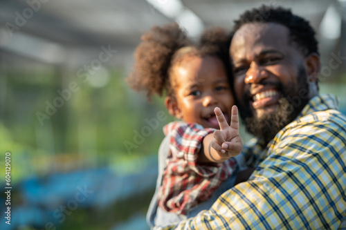 Happy african black parents dad father with daughter child on piggyback teasing fun in garden greenhouse. Black daughter kiss cheek dad and neck riding in vegetable greenhouse garden in light sunset © BESTIMAGE