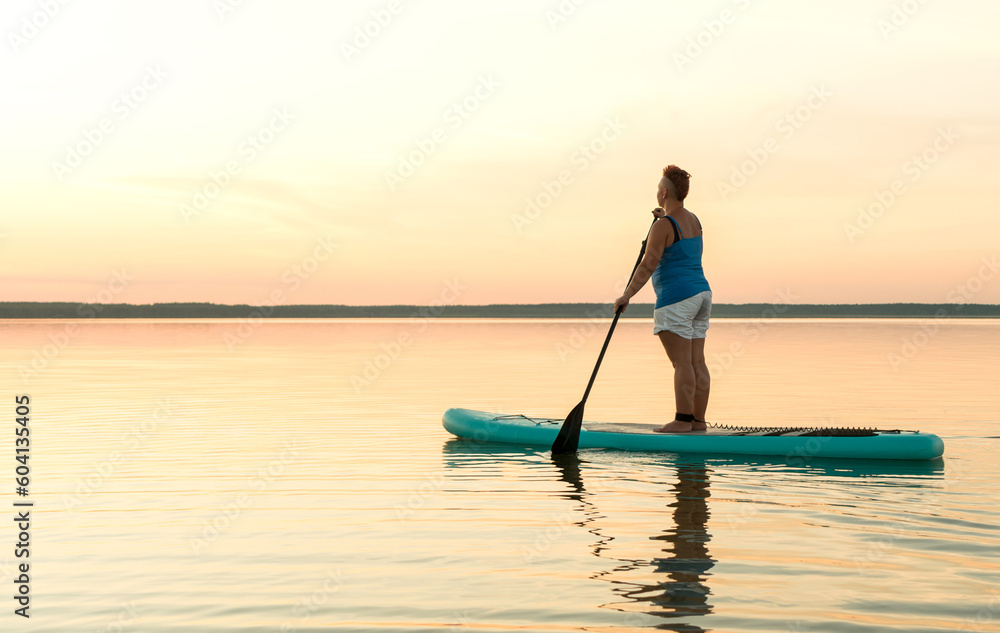 A woman in mohawk shorts stands on a SUP board at sunset in a lake against a pink-blue sky and water.