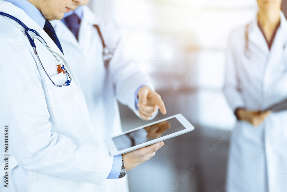 Group of unknown doctors use a computer tablet to check up some medical names records, while standing in a sunny hospital office. Physicians ready to examine and help patients. Medical help, insuranc Group of unknown doctors use a computer tablet to check up some medical names records, while standing in a sunny hospital office. Physicians ready to examine and help patients. Medical help, insuranc