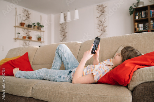A teenage girl lies on a sofa and plays or chats with friends using a mobile phone. Children use gadgets in everyday life.