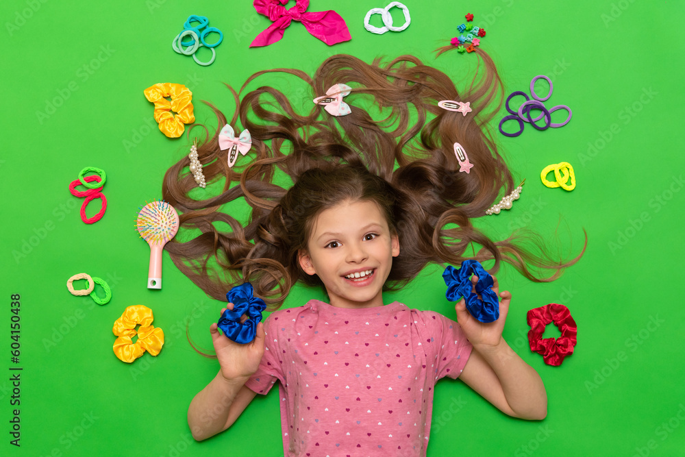 A little girl lies among the hair bands and hairpins. A child with loose hair and hair accessories is lying on a green isolated background.